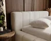 A close-up view of a bed with a textured boucle headboard and white bedding, set against a wood-paneled wall. Náhľad obrázku produktu