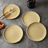 Four plates are arranged on a gray surface, with cutlery on one plate and a vase in the background. Produktbild-Vorschau