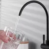 A hand is holding a glass while water is being poured into it from a black faucet. Produktbild-Vorschau