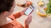 A person is holding a smartphone displaying a recipe while standing in a kitchen with ingredients for baking. Produktbild-Vorschau