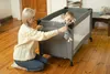 A woman is kneeling next to a travel crib, playing with a baby inside. Anteprima dell’immagine prodotto