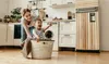 A mother and child are playing in a laundry room, with the child sitting in a laundry basket. Produktbild-Vorschau