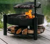 A black metal fire pit with a grill rack cooking sausages over a fire, set on a wooden deck next to a body of water and greenery. Náhled obrázku produktu
