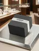 A dark gray watch box with a silver band is placed on a metallic surface in a jewelry store. Voir les images du produit