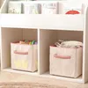 A white bookshelf with two storage bins on the bottom shelf. The left bin has a crown design and contains colorful toys. The right bin is textured and contains folded clothes. Podgląd zdjęcia produktu