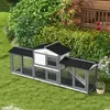 A multi-level wooden hutch for small animals, set up in a garden with lush green bushes and a wooden building in the background. Produktbild-Vorschau