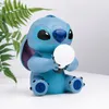 A blue Stitch figurine holding a white ball, sitting on a white surface with a green fern in the background. Produktbild-Vorschau