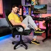 A man is sitting in a gaming chair in a room with a computer setup, talking on the phone. Produktbild-Vorschau