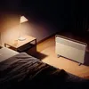 A bedroom scene at night with a heater, a bedside table with a lamp and a glass of water, and a bed. Produktbild-Vorschau