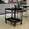 A black metal serving cart with wheels is shown in a cafe or restaurant setting. Náhled obrázku produktu