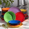 A collection of colorful ceramic plates and bowls stacked and arranged on a white surface, with greenery and candle holders in the background. Náhled obrázku produktu