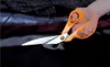 A hand holding a pair of orange-handled scissors, about to cut black fabric. Podgląd zdjęcia produktu
