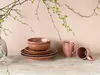 A stack of brown ceramic bowls and plates, along with two pink ceramic mugs, are arranged on a wooden surface. Delicate branches with green leaves are draped from above, creating a natural and elegant setting. Náhľad obrázku produktu