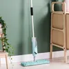 A spray mop is shown standing on a wooden floor in front of a green wall, with a small wooden stool and a plant to the left. Náhled obrázku produktu