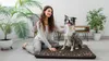 A woman is sitting on the floor next to her dog, which is lying on a dog bed. They are in a well-lit room with plants and furniture. Náhled obrázku produktu
