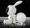 A white astronaut-like figure with rabbit ears sits on a textured surface resembling the moon, next to a sphere that looks like the moon. Náhled obrázku produktu