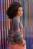 A woman is posing in front of a colorful striped wall, wearing a sparkly sweater. Produktbild-Vorschau