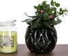 A black ceramic planter with a diamond pattern and rhinestones, containing a green plant with small pink flowers, sits next to a lit green candle in a glass jar. Produktbild-Vorschau