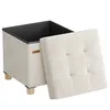 A cream-colored storage ottoman with its lid open, revealing a black interior. The ottoman has wooden legs and a small brown tag on the side. Produktbild-Vorschau