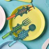 A set of children's dinosaur-themed cutlery is arranged on a yellow plate, with other plates visible in the background. Produktbild-Vorschau