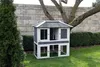 A two-story wooden rabbit hutch is set up on a green lawn in front of a stone wall and a large tree. Voir les images du produit