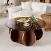 A round coffee table with a glass top is placed in a living room setting, next to a white sofa. Decorative items like fruit and flowers are on the table. Produktbild-Vorschau