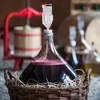 A close-up of a glass carboy filled with red wine, with a fermentation airlock on top. The carboy sits in a basket lined with straw. In the background, a red fruit press and barrels are visible. Produktbild-Vorschau