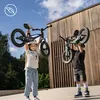 Two children are holding up balance bikes outdoors in front of a wooden building. Produktbild-Vorschau