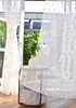 A sheer curtain with gold pom-poms hangs in front of a window with plants visible outside. A small side table with a vase and a basket is placed in front of the curtain. Produktbild-Vorschau