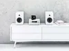 A white stereo system with two speakers, a receiver, and a small black device sits on a white cabinet against a textured gray wall. Anteprima dell’immagine prodotto