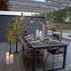 An outdoor dining table set up on a wooden deck under a pergola, decorated with string lights and candles. Produktbild-Vorschau