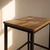 A close-up of a bar stool with a wooden top and black metal legs, with light casting shadows on the wall behind it. Podgląd zdjęcia produktu