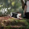 A mobile hotspot sits on a moss-covered log in a forest setting, with a person blurred in the background. Produktbild-Vorschau