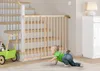 A baby is crawling on the floor near a wooden baby gate installed at the bottom of a staircase. A toy stroller is also visible. Produktbild-Vorschau
