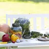 A table set up outdoors with jars of pickles and fresh vegetables, suggesting a home canning or preserving setup. Produktbild-Vorschau