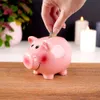 A hand is inserting a coin into a pink ceramic piggy bank. There are lit candles in the background. Náhľad obrázku produktu