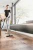 A woman is vacuuming a spill on a wooden floor in a modern kitchen with large windows. Náhled obrázku produktu
