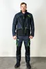 A man is standing and posing in front of a white background, wearing a blue and black work suit with green accents. Produktbild-Vorschau