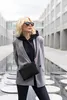A woman wearing a grey blazer and black hoodie stands in an outdoor urban setting with a reflective pool in front of her. Produktbild-Vorschau