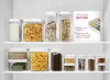 A pantry shelf filled with various clear storage containers holding dry goods like pasta, cereal, crackers, and spices. Anteprima dell’immagine prodotto