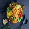 A vibrant and colorful salad bowl with grilled cheese, couscous, chickpeas, and fresh vegetables, presented on a dark background with serving utensils. Produktbild-Vorschau