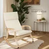 A cream-colored rocking chair is placed on a fluffy rug in a well-lit room, with a large plant and a side table with a lamp in the background. Náhled obrázku produktu