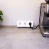 A white multi-socket power strip with a black plug is shown on a grey countertop next to a coffee machine. Produktbild-Vorschau