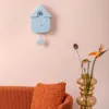 A light blue cuckoo clock hangs on a pink wall above an orange couch with an orange textured pillow. To the right, a vase holds a bird of paradise flower and other greenery. Náhled obrázku produktu