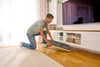 A man is kneeling on the floor in a living room, assembling or adjusting a baby bouncer. Produktbild-Vorschau