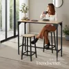 A woman sits at a bar table, holding a mug, in a modern interior setting. Náhled obrázku produktu