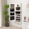 A white jewelry cabinet with its doors open, displaying jewelry and makeup, is mounted on a textured wall next to a large potted plant and a white nightstand with a decorative sculpture. Náhľad obrázku produktu