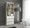 A tall, narrow bookshelf with white drawers and light wood shelves, standing against a textured gray wall. A floor lamp is to the right, and a white sofa is partially visible in the foreground. Produktbild-Vorschau