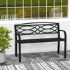 A black metal garden bench is shown on a stone patio with a rug in front of it. A potted plant is to the left of the bench. A window with reflections of grass and objects is behind the bench. Podgląd zdjęcia produktu