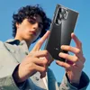 A young man is holding a clear phone case with a Samsung phone inside against a bright blue sky. Produktbild-Vorschau